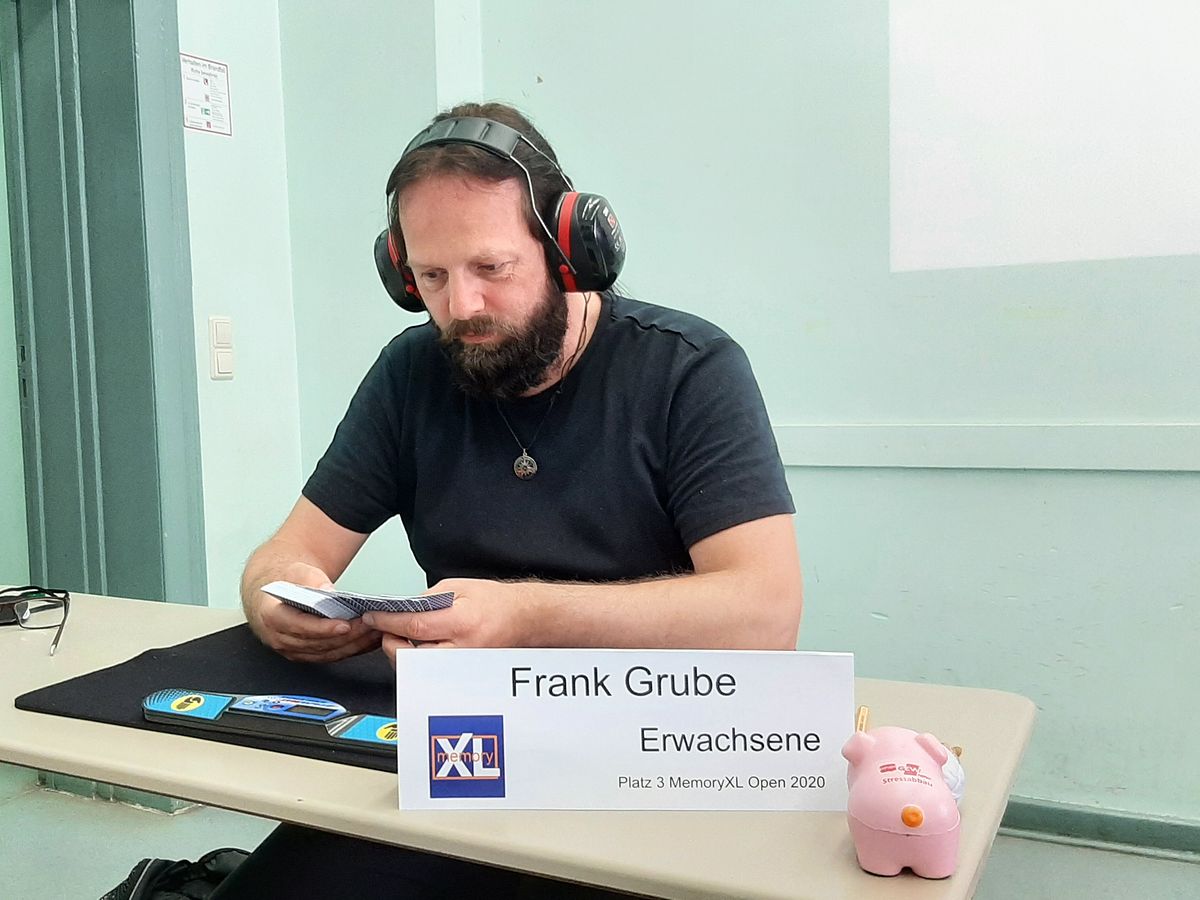 Frank Grube mit rotem Gehörschutz prägt sich einen Kartenstapel ein. Vor ihm steht das Namensschild „Frank Grube, Erwachsene, Platz 3 MemoryXL Open 2020“.